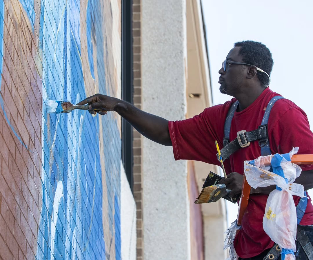 Adeodunfa painting a mural on site, brush in hand, working over blue on a brick wall.